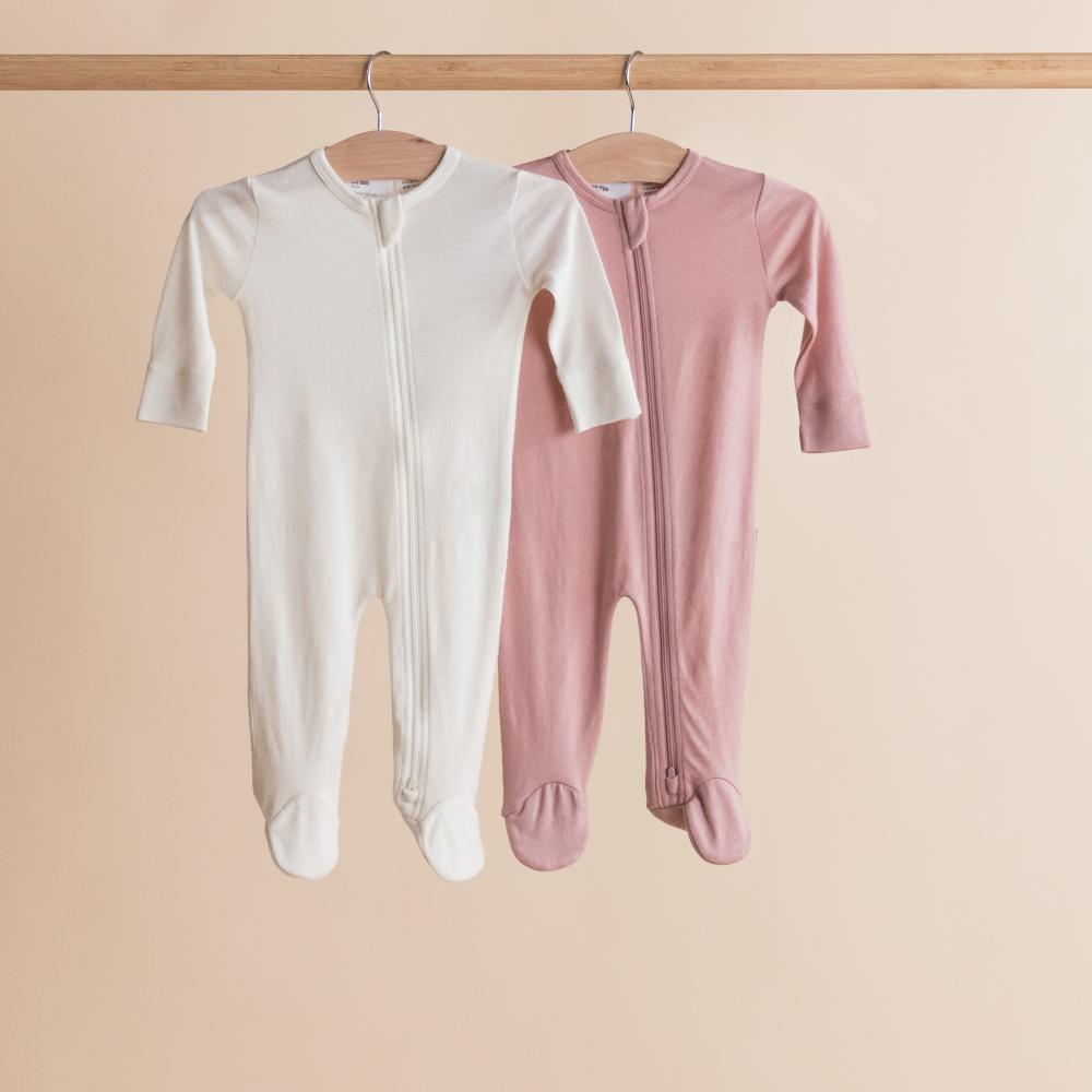 Two merino baby onesies, one white and one pink, hanging on a wooden rod against a beige background.