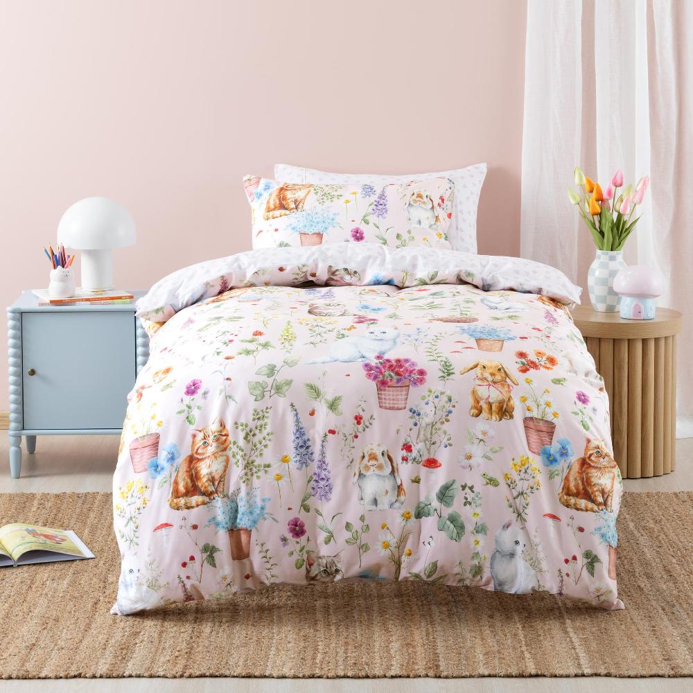 A pastel bedding set featuring a whimsical tea-and-cats print with illustrated cats, teacups, flowers, and mushrooms on a light pink background, styled in a bedroom setting with a nightstand, lamp, and flowers.