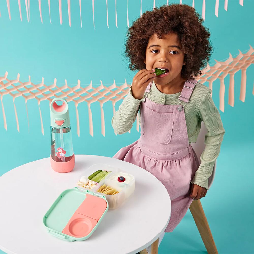 Child in pink overalls eating from a lunchbox with a water bottle on a teal background
