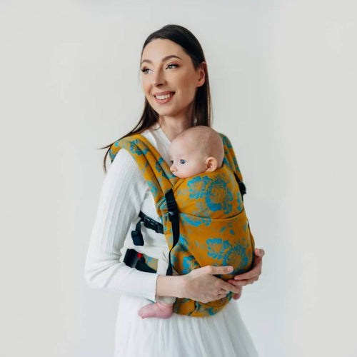 A woman holding her baby in a baby carrier. The carrier is a mustard colour with a teal green floral pattern. 