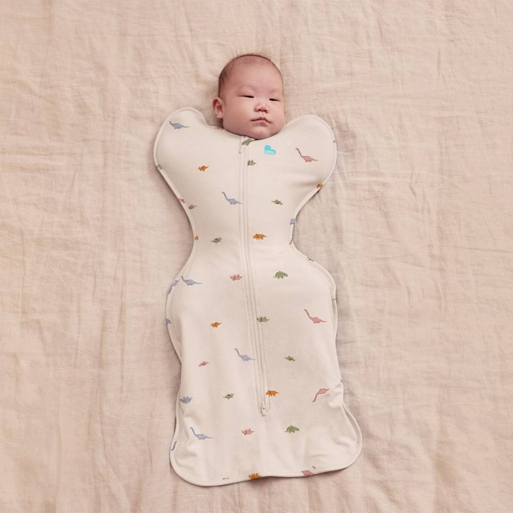 A sleeping baby lying on their back on a textured beige linen blanket, wearing a baby swaddle with a zipper and a small multicolor dinosaur print.