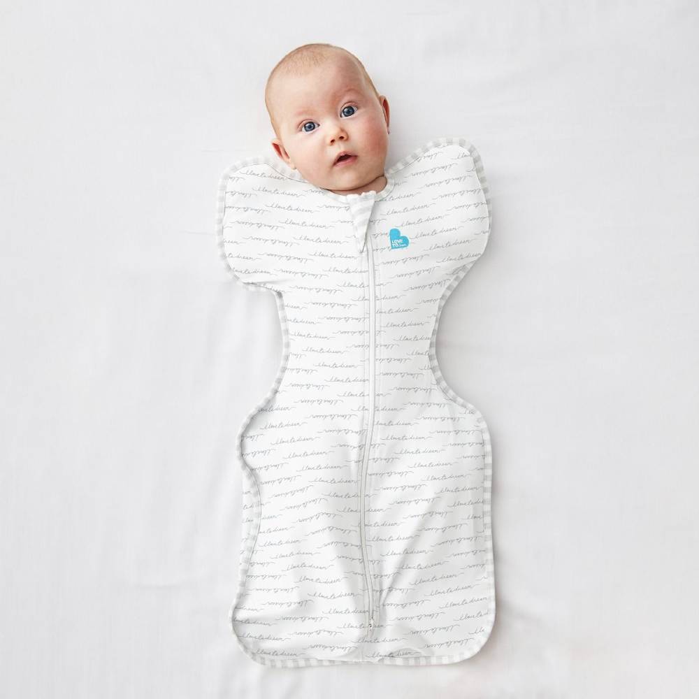 A baby lying on a white blanket, wrapped in a white zippered swaddle blanket with a light gray script pattern and a small light blue heart logo.