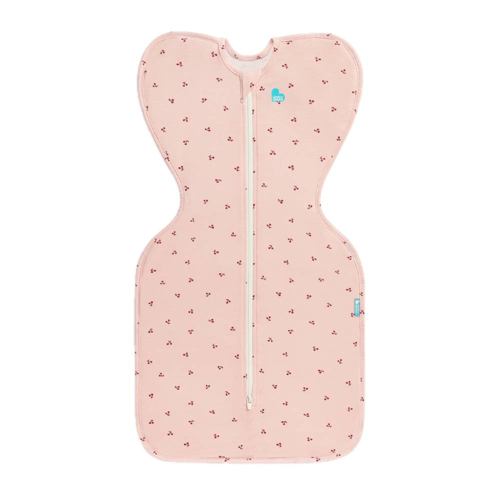 A light pink baby sleep swaddle with a small repeating red cherries pattern, featuring a central zipper and a small blue logo on the upper right side, shown against a plain white background.