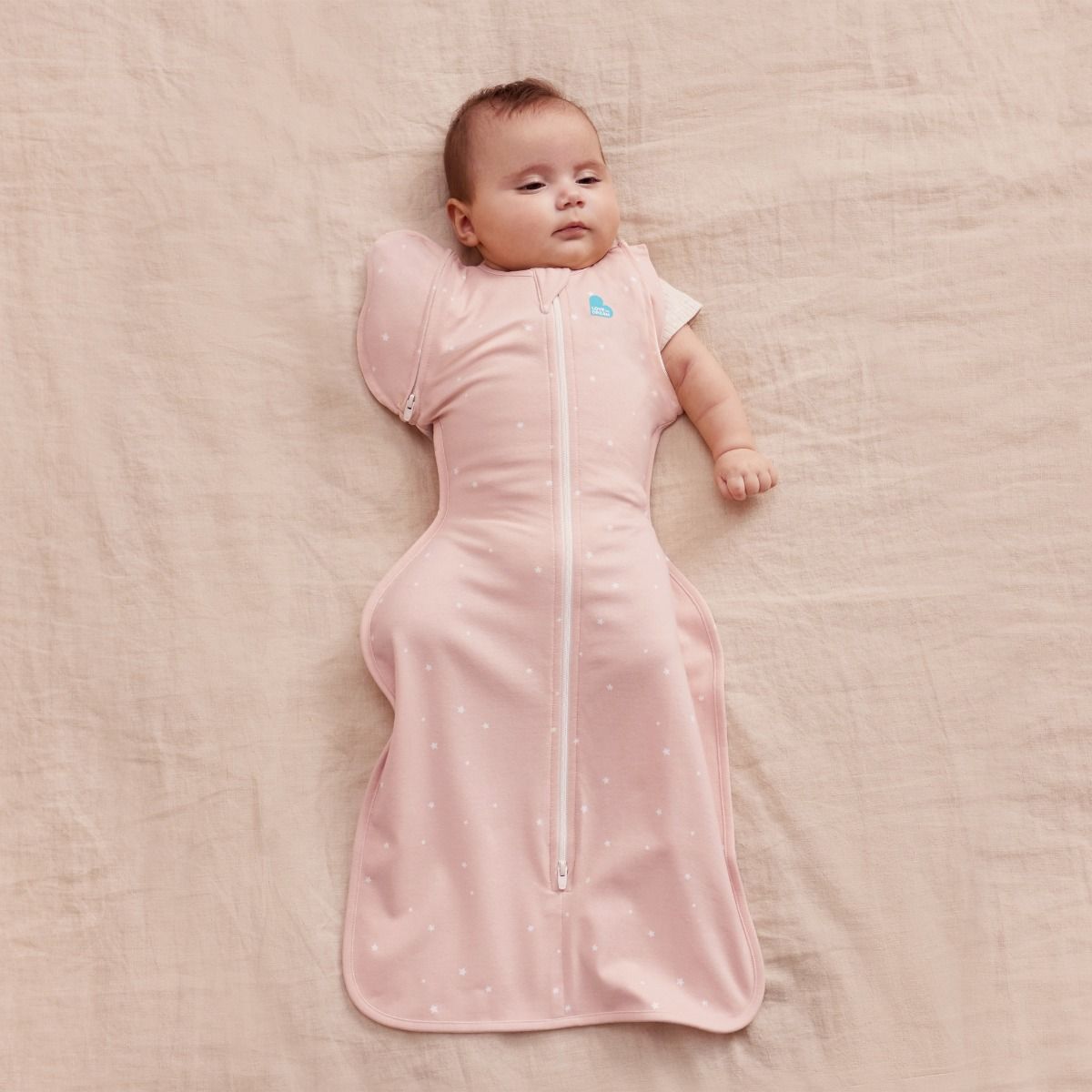 A baby lying down wearing a 1.0 tog Swaddle Up Transition Bag - Powder Pink Twinkle with one arm out. 