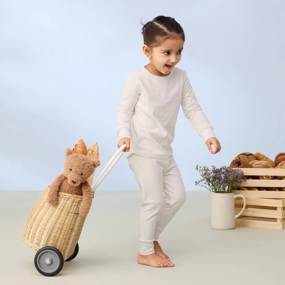 Child in beige pyjamas pushing a woven cart with a plush toy, on a light background
