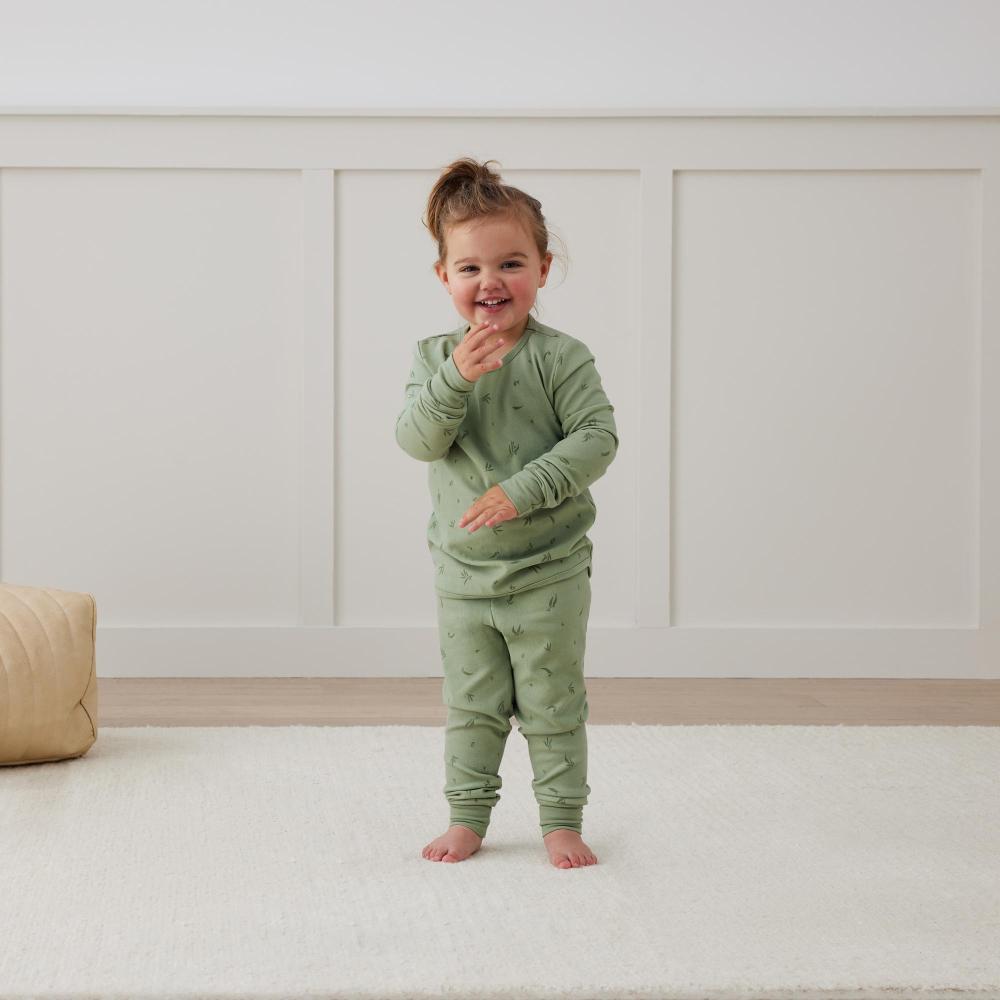 Child wearing green pajamas standing on a white floor with a neutral-colored wall in the background
