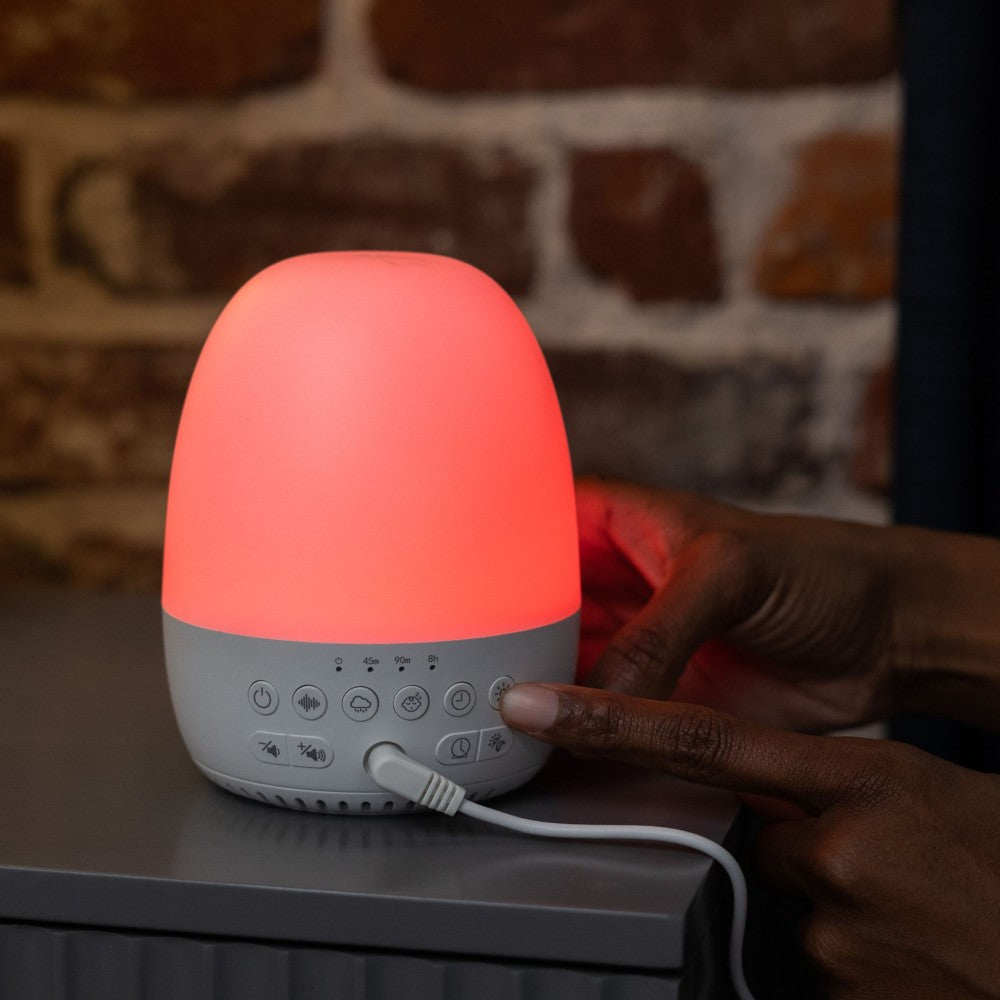 Sleep trainer clock with a glowing red light on a surface, with a person's hand interacting with it against a brick wall background.
