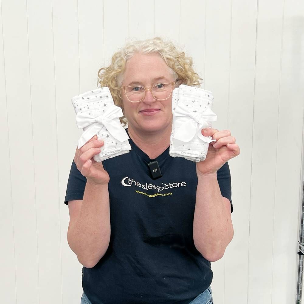 A woman with curly blonde hair and glasses holds up two white baby wraps or cloths with small dark stars and white bows, wearing a navy blue t-shirt with 'thesleepstore' printed on the front, against a white paneled wall background.
