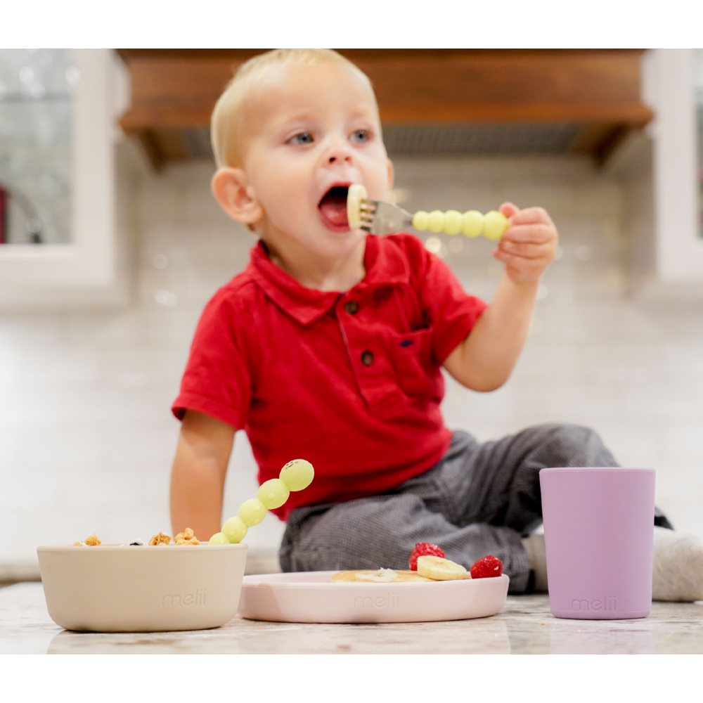 Child in a red shirt eating with a yellow fork, sitting at a table with a bowl and cup.