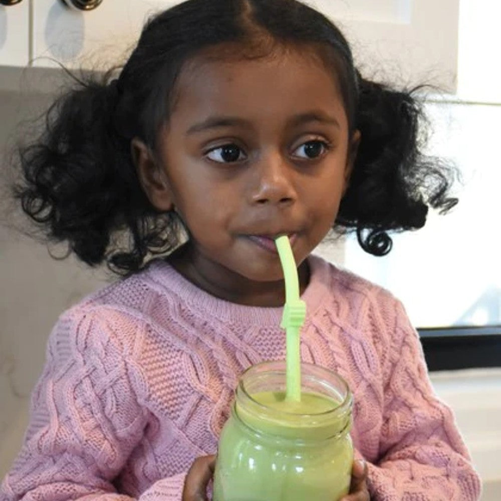 Child holding a green smoothie with a straw in a kitchen setting