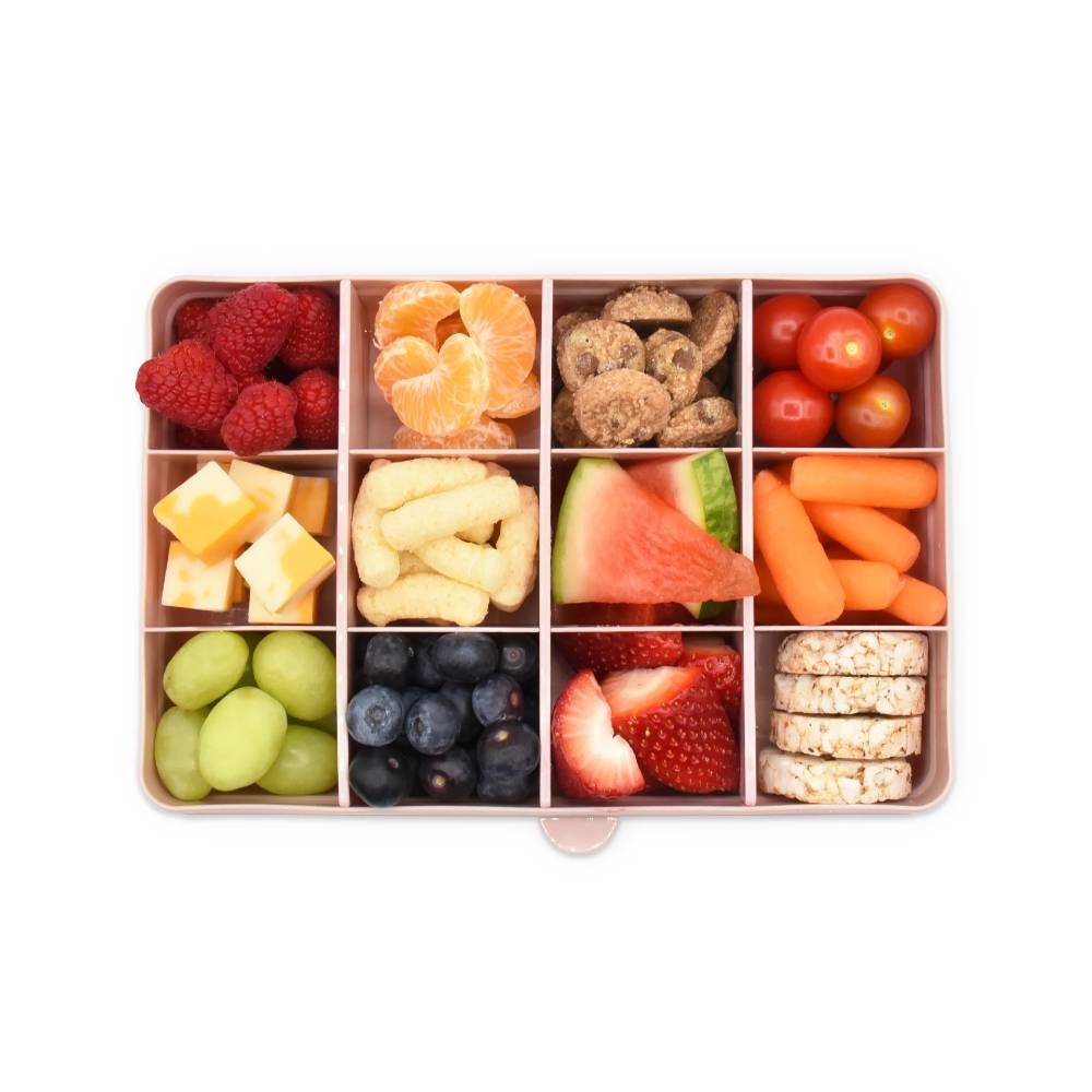 Assorted fruits, vegetables, and snacks in a compartmentalized container on a white background