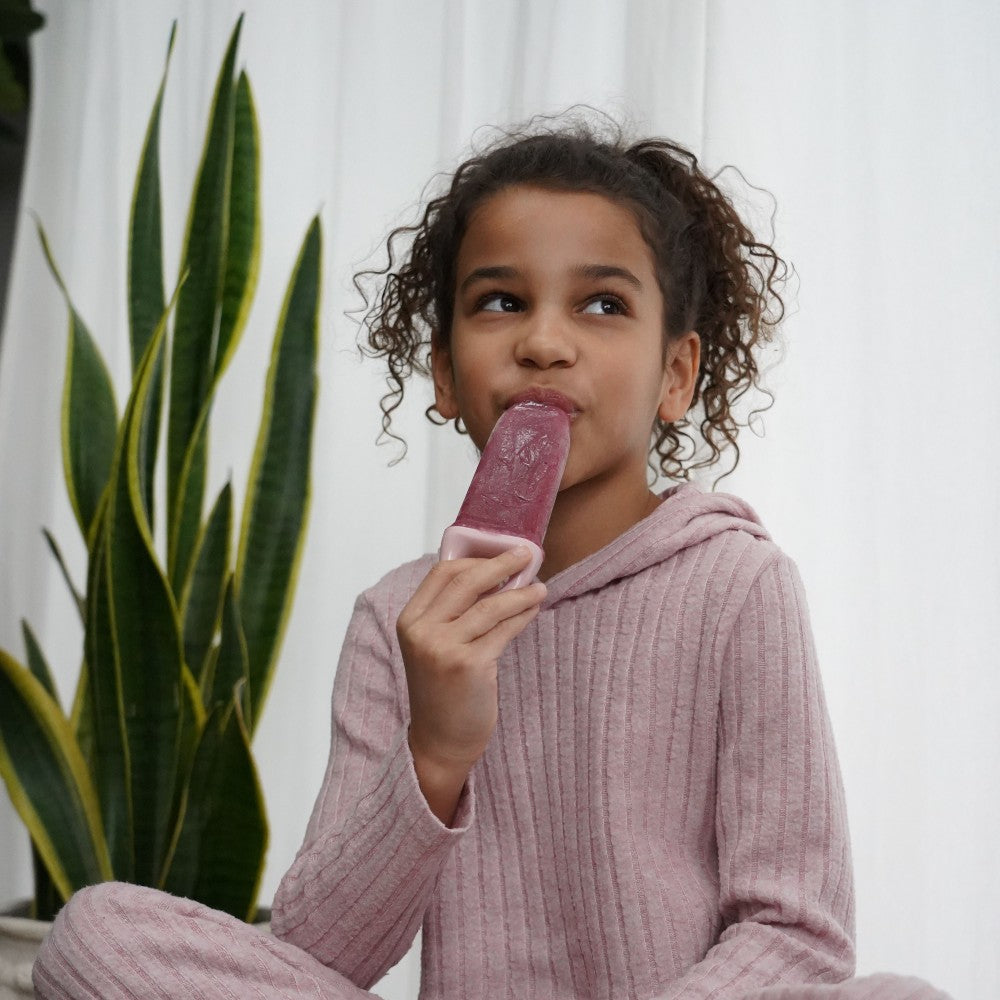 Child in pink pajamas eating a popsicle with a plant in the background