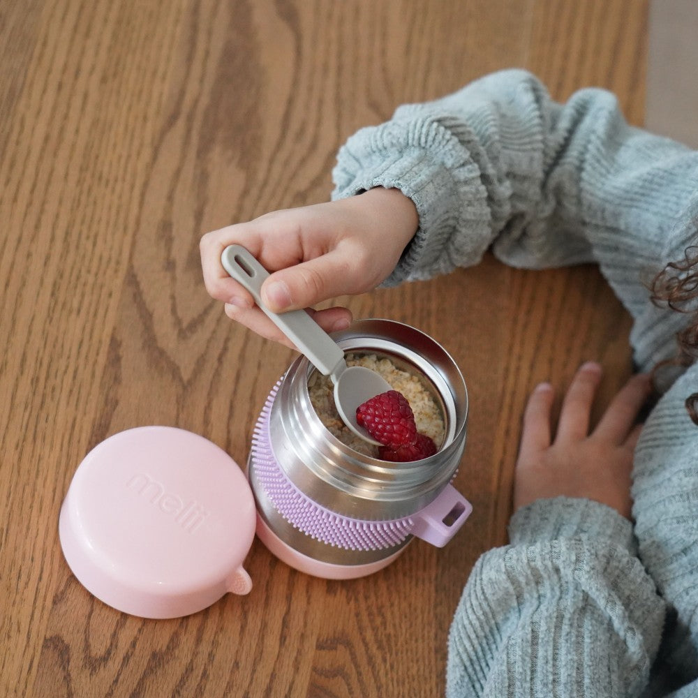 Child's hand using a spoon to eat from a pink and silver thermos on a wooden surface