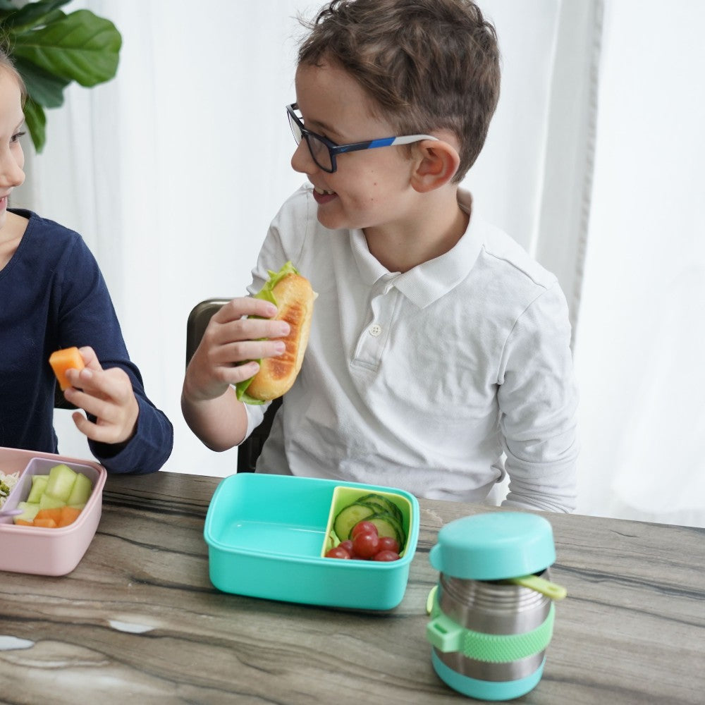 Two children eating at a table with a teal lunch box and container.