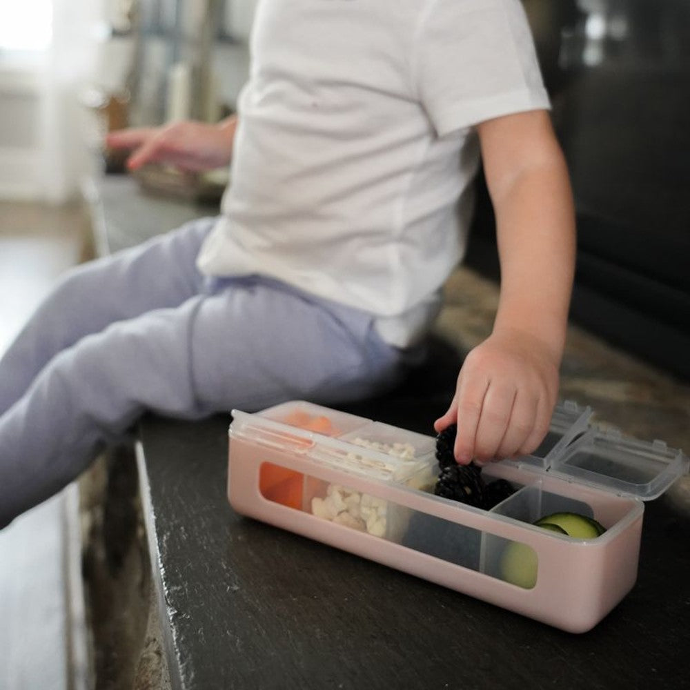 Child reaching into a pink Melii Snackle bento box on a kitchen counter
