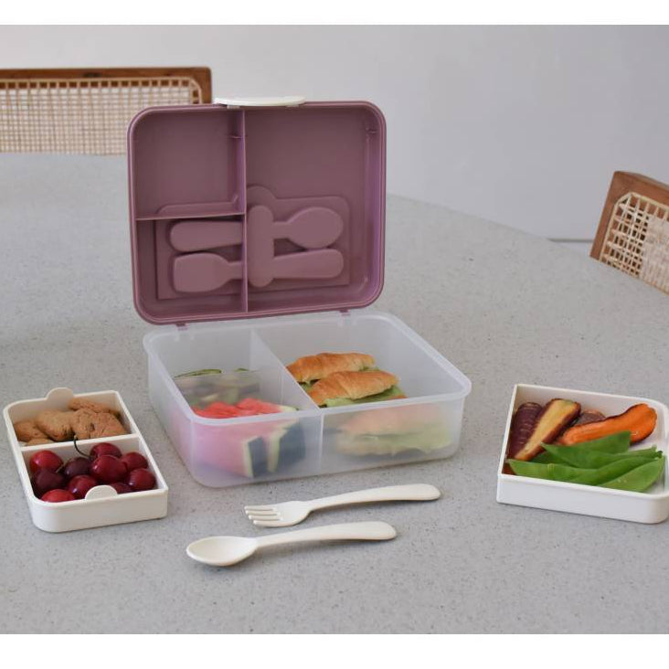 Bento box with compartments containing food on a table with chairs and a plant.