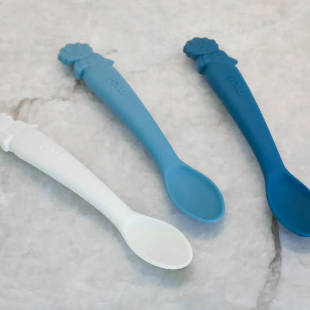 Two blue and one white silicone Melii baby spoon on a textured surface