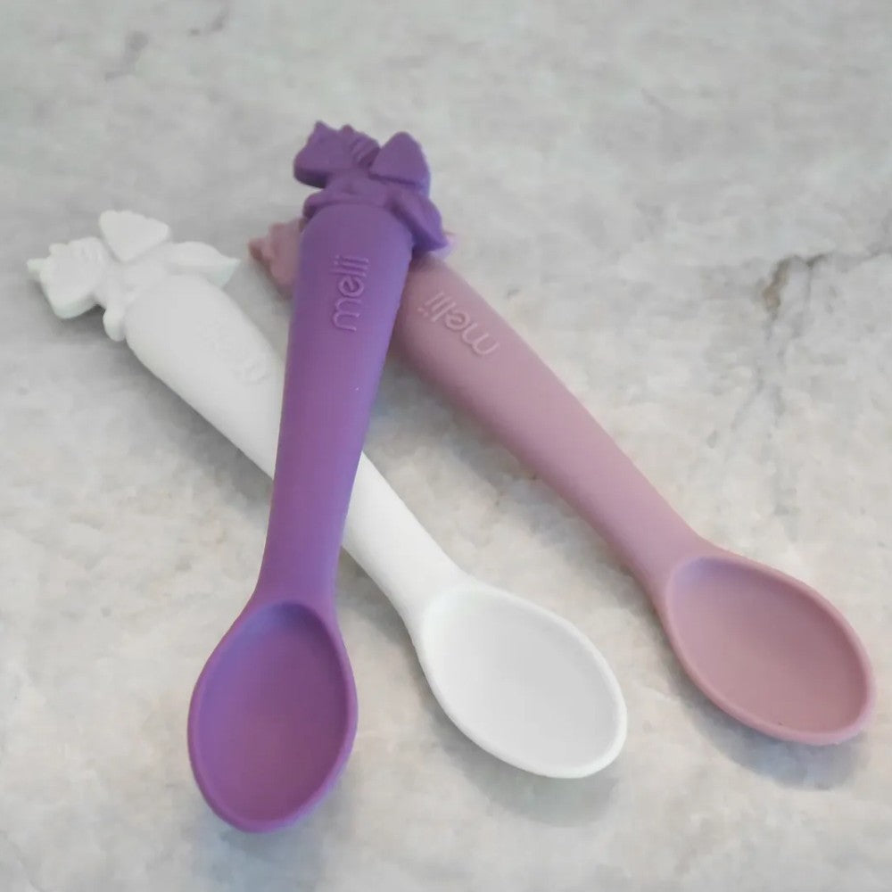 Two silicone spoons, one purple and one pink, on a textured surface.