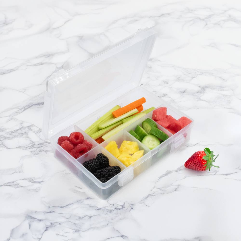 Transparent snack container with compartments filled with fruits and vegetables on a marble surface