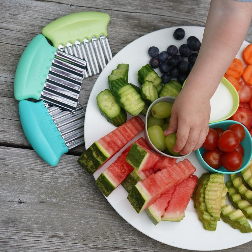 Assorted fruits and vegetables on a plate with a child's hand reaching for grapes, surrounded by kitchen tools.
