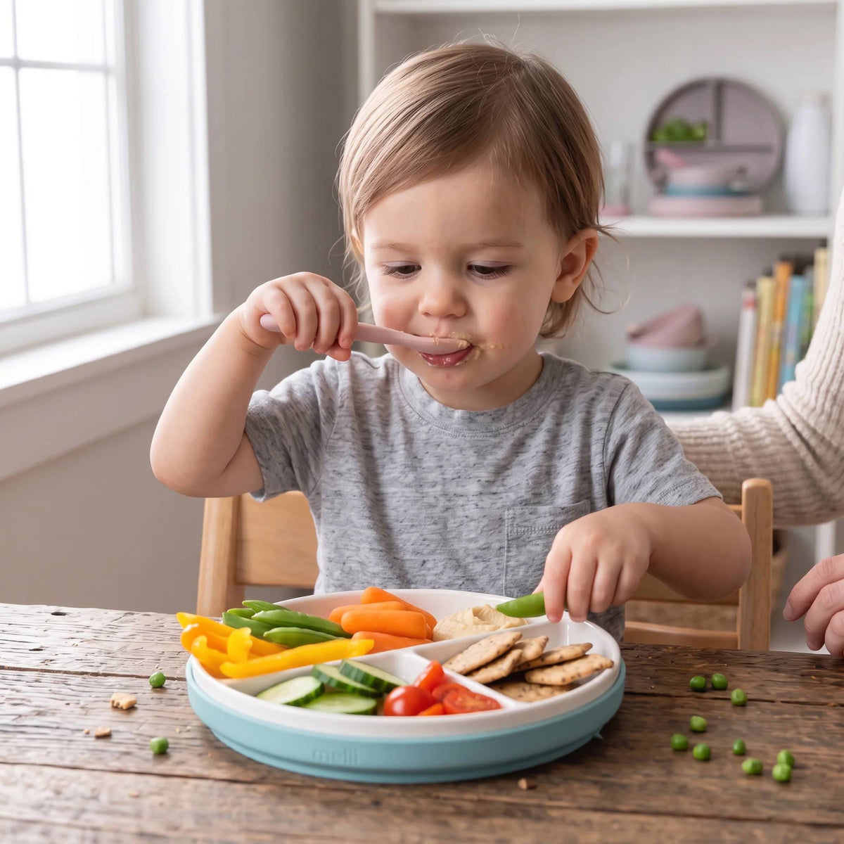 A young child eating a selection of foods from a divided ceramic plate with a blue silicone sleeve. 