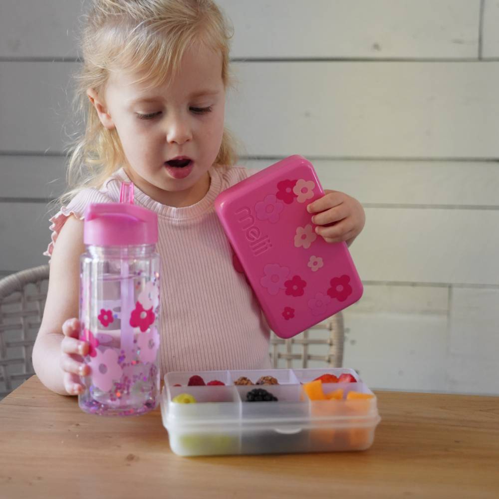 Child holding a pink lunchbox, water bottle, and snack container with colorful snacks.