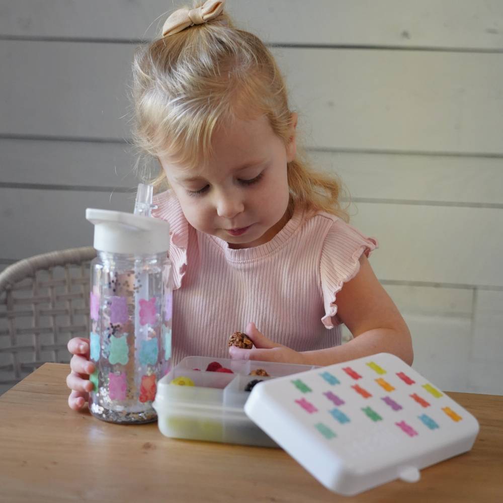 Child playing with a colorful snack container and water bottle on a wooden table.