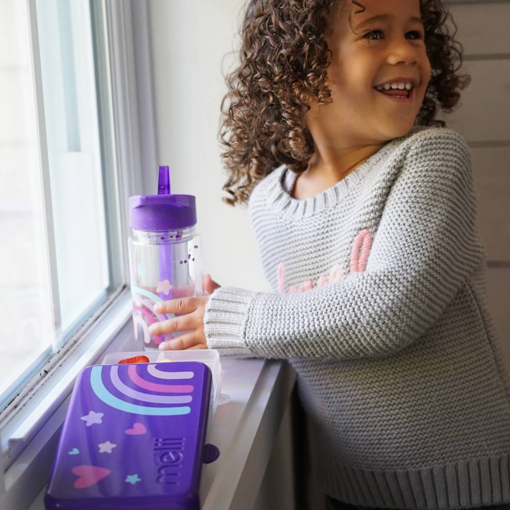 Child with curly hair wearing a gray sweater, standing by a window with a purple tumbler and lunchbox.