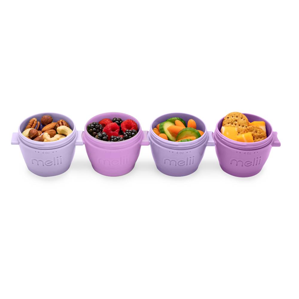 Four small round plastic snack bowls in light purple, arranged in a row on a white background. Each bowl is filled with different foods: nuts, mixed berries, sliced fruit, and crackers.