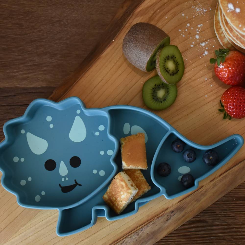Blue dinosaur-shaped plate with snacks on a wooden table with fruits