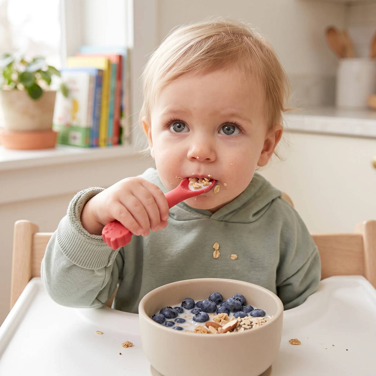 A young child eating cereal and blueberries with a red strawberry theme silicone spoon. 
