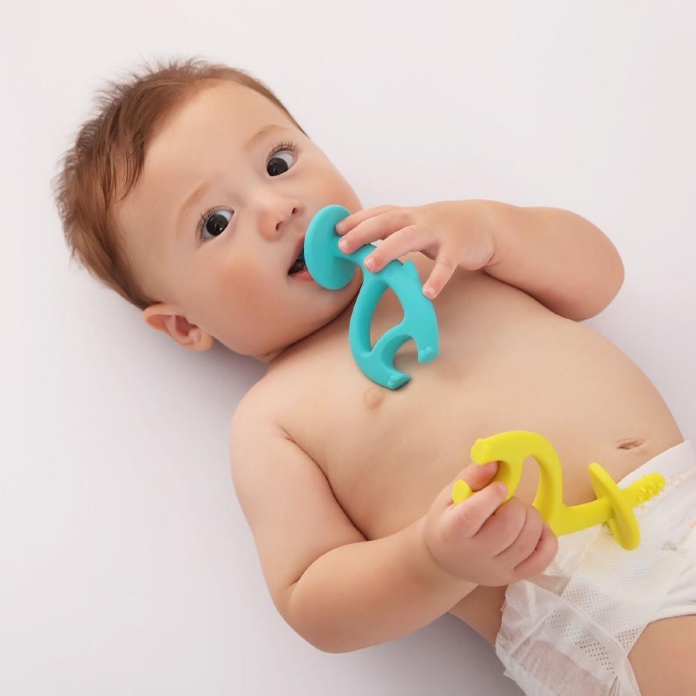 A baby lying on their back holding a turquoise pacifier clip in their mouth and another yellow pacifier clip in their hand, wearing a white diaper against a plain white background.