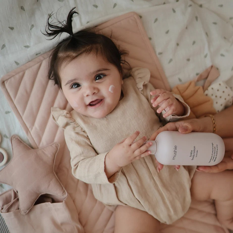 A baby wearing a beige long-sleeve romper lies on a pink quilted blanket while an adult holds a white bottle of mushie baby lotion near the baby's face.