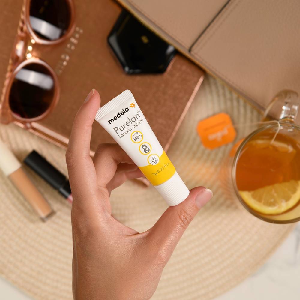 A hand holds a tube of Medela Purdlar Lotion Cream against a styled flat-lay background with rose gold sunglasses, a glass of tea with a lemon slice, and other cosmetics.