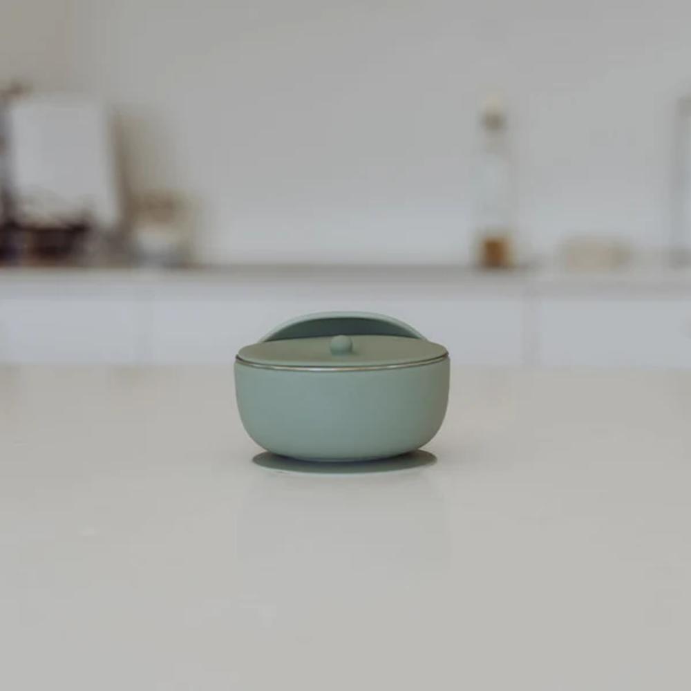 A small round silicone lidded container in a pale sage green color, set on a light gray surface with a blurred, minimalist background.