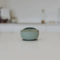 A small round silicone lidded container in a pale sage green color, set on a light gray surface with a blurred, minimalist background.