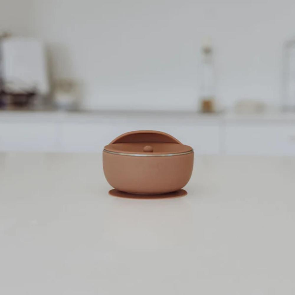 A small terracotta-colored ceramic lidded container with a rounded dome lid and a tiny central knob, sitting on a light gray surface with a blurred, minimalist background.