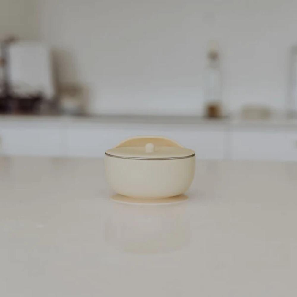 A small cream-colored silicone and stainless steel bowl with a matching lid,  set on a light-colored countertop with a blurred kitchen background.