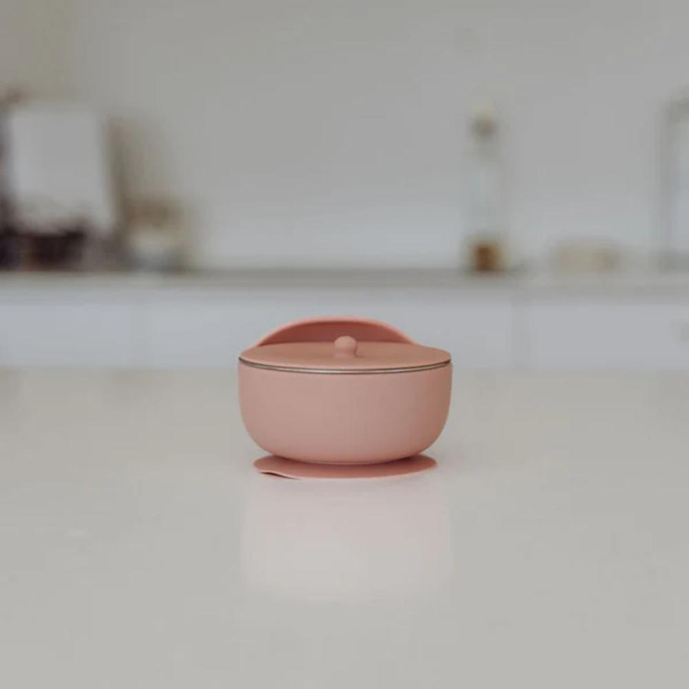 A small dusty pink silicone lidded bowl with suction base set on a clean white surface with a blurred kitchen background.
