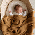 A sleeping newborn baby in a white ribbed long-sleeve outfit, nestled inside a woven natural fiber bassinet and wrapped in a textured brown blanket.