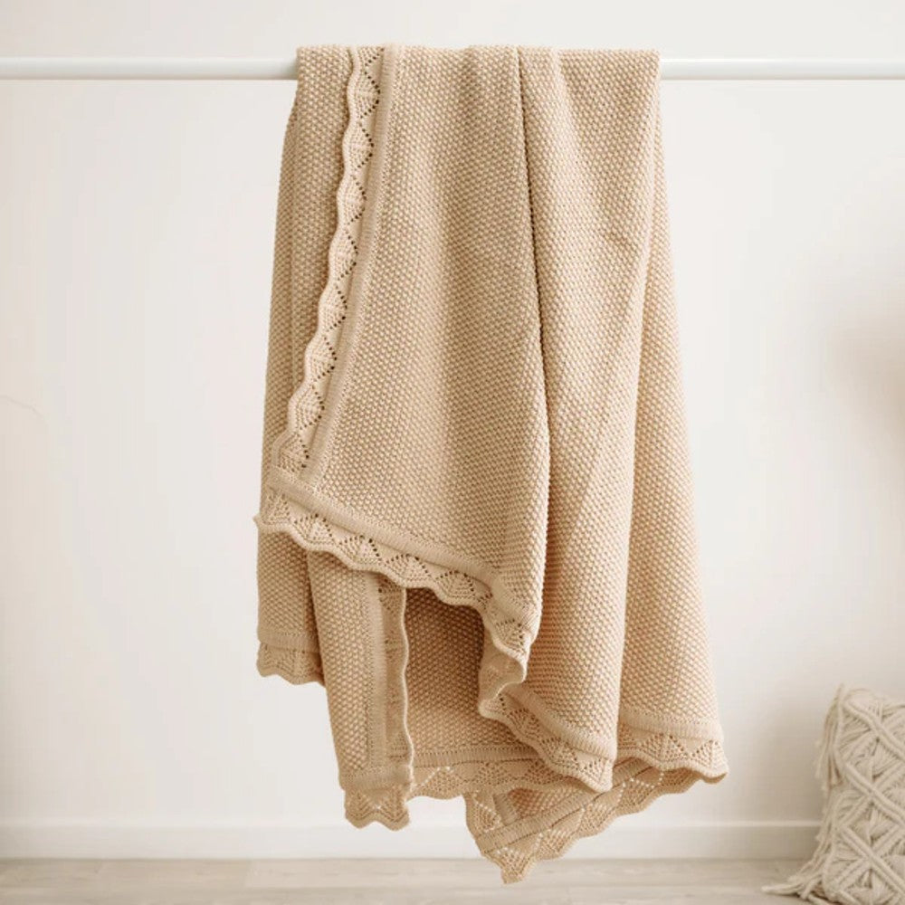 A beige textured throw blanket with scalloped edge trim hanging from a white rod against a white wall.