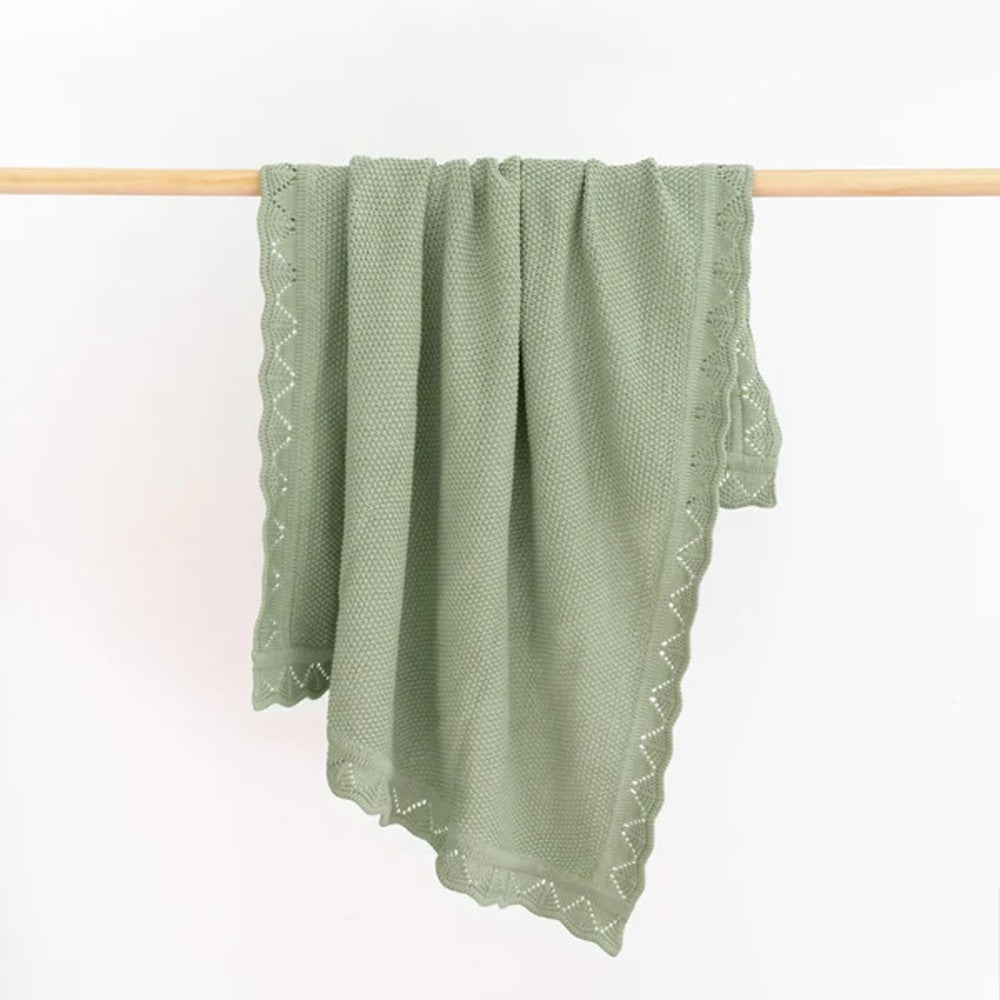 A sage green textured throw blanket with a scalloped embroidered border hangs from a light wood dowel against a plain white background.