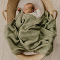 A newborn baby wrapped in an olive green textured knit blanket, lying inside a woven straw Moses basket on a light wood floor.