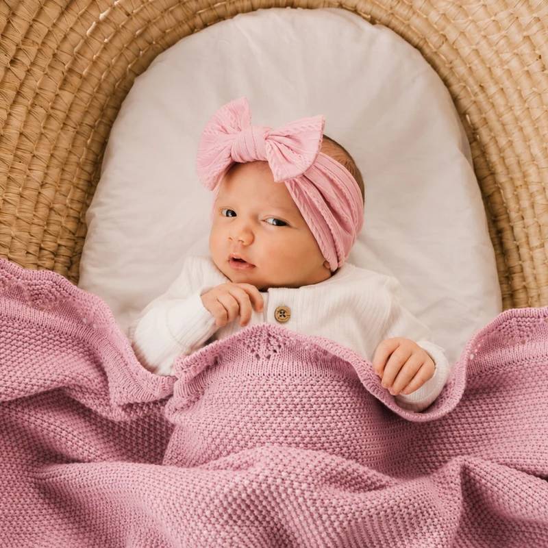 Baby in a crib wearing a pink headband and surrounded by a pink blanket.