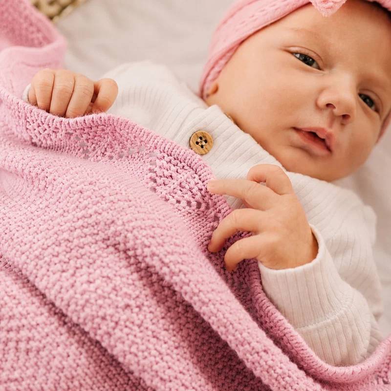 A close-up of a baby wearing a white ribbed knit top and pink hat, wrapped in a chunky pink knit blanket.