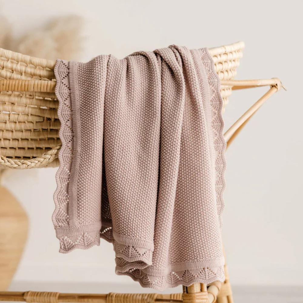 A soft knit baby blanket in a dusty mauve-pink color with scalloped lace trim, draped over a woven rattan chair.