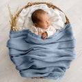 A sleeping newborn baby wrapped in a textured dusty blue blanket, resting inside a woven straw basket lined with a white dotted blanket on a light gray background.