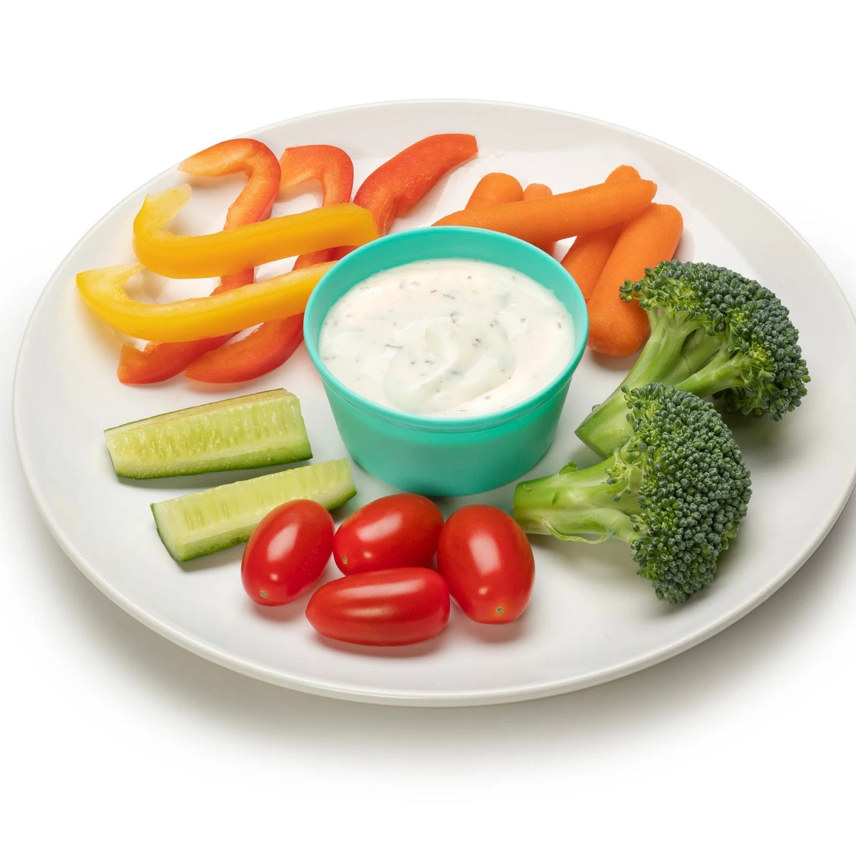 A white plate arranged with a small turquoise cup of white dipping sauce, surrounded by baby carrots, broccoli florets, cucumber sticks, cherry tomatoes, and sliced red and yellow bell peppers on a white background.