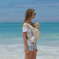 A woman at the beach holding her baby in a Mini Monkey baby carrier in the colour sand. 