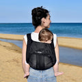 A woman at the beach holding her baby on her back in a Mini Monkey Toddler carrier in the colour black. 
